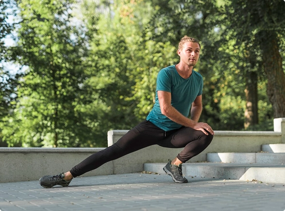 A man in athletic wear performs a lateral lunge stretch outdoors on a paved path with trees and stairs in the background.