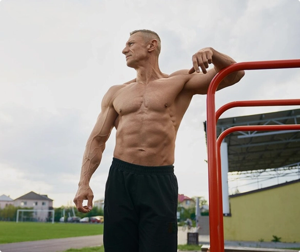 A shirtless man stands confidently on a red rail, showcasing a relaxed posture against a neutral background.