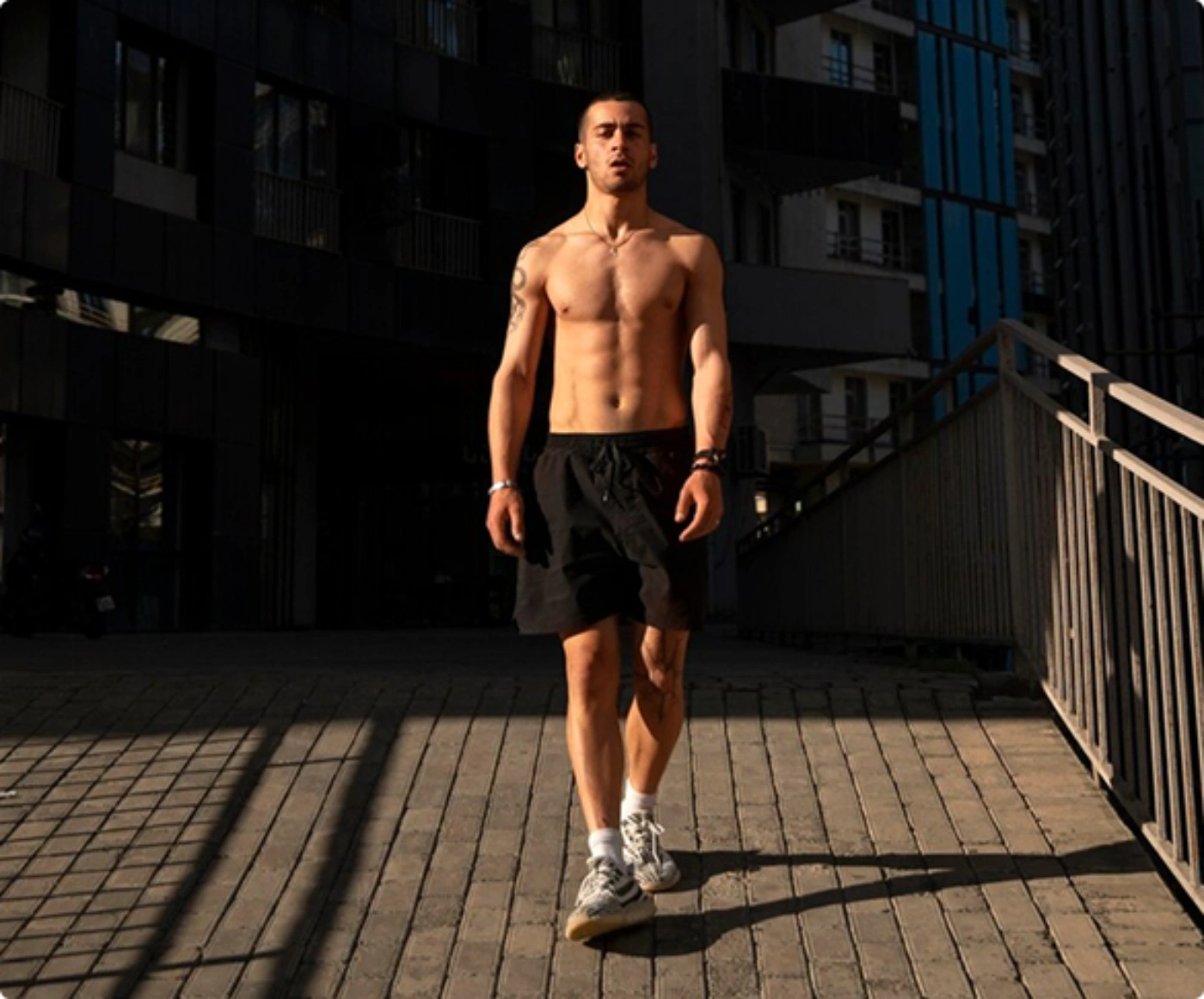 A shirtless man walks on a sidewalk, with tall buildings towering in the background.