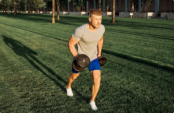 A man exercises on a grassy field, running with two dumbbells in his grip.