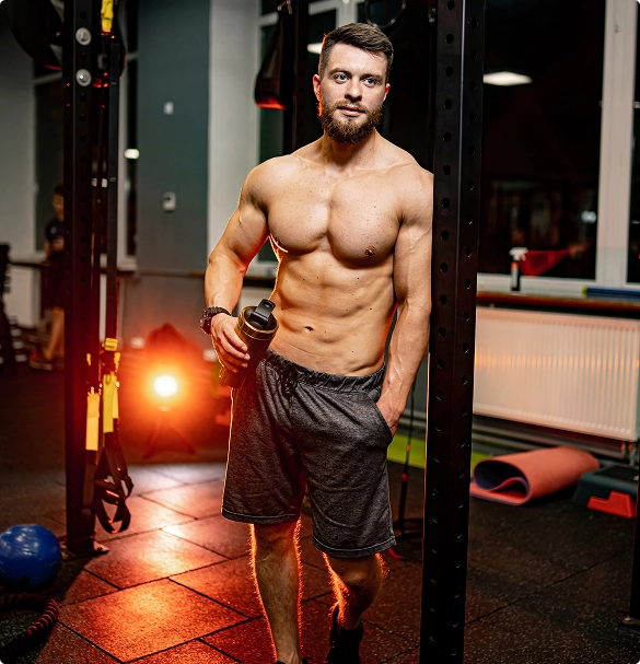 A shirtless man is seen in a gym, standing near fitness equipment, emphasizing a focus on physical training.