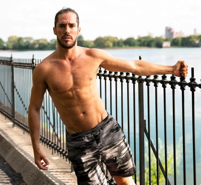 A shirtless man leans against a fence, overlooking a tranquil body of water in a serene outdoor setting.