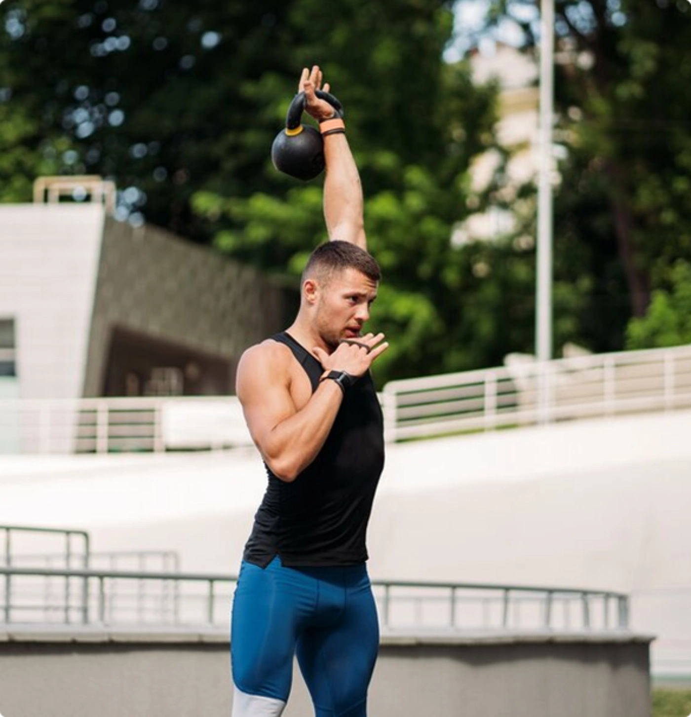 A man performs a kettlebell exercise in a park, focusing on strength training amidst greenery.
