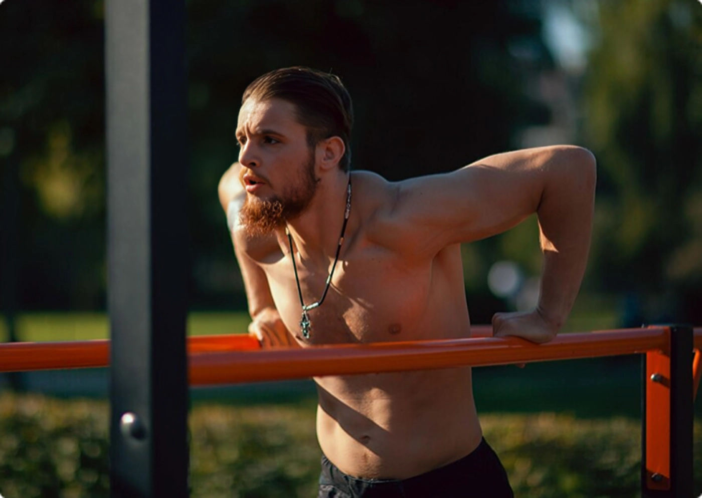 A shirtless man performs pull-ups on a bar, showcasing strength and fitness in an outdoor setting.