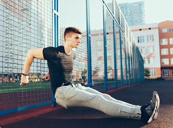 Young man in athletic wear doing tricep dips against a blue fence on an outdoor track. The scene conveys focus and determination in a sunny setting.