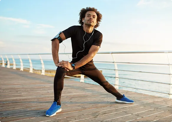 Man in athletic gear stretches on a boardwalk by the sea. He wears headphones and a phone armband, exuding focus and energy under a clear sky.