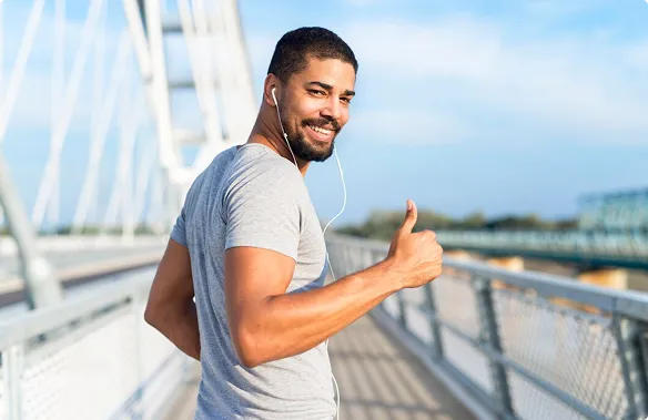Man in a gray t-shirt with earbuds gives a thumbs-up, smiling while standing on a bridge. He exudes confidence and positivity under a clear blue sky.