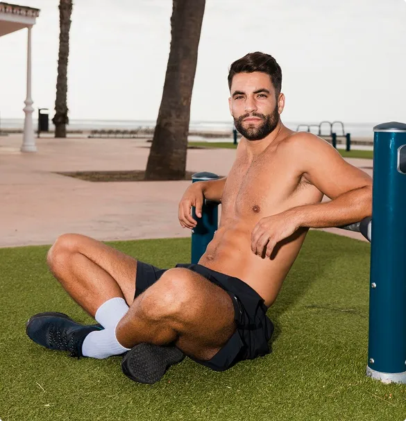 Shirtless man sitting on grass in a park near palm trees, leaning against a blue pole. He wears black shorts, white socks, and black shoes, looking relaxed.