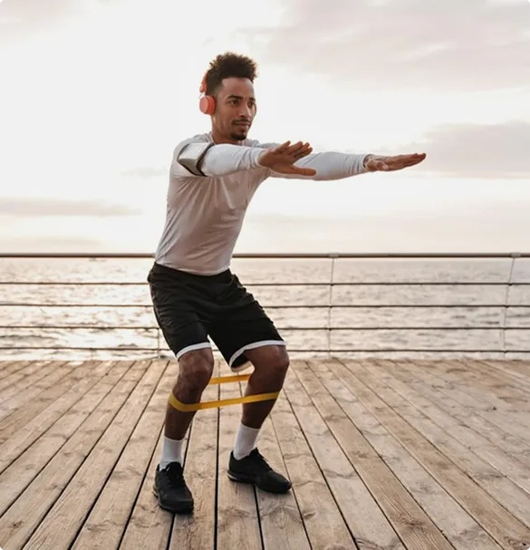 A man exercises with a resistance band by the ocean at sunset. He wears headphones, a long-sleeve shirt, and shorts, conveying focus and energy.