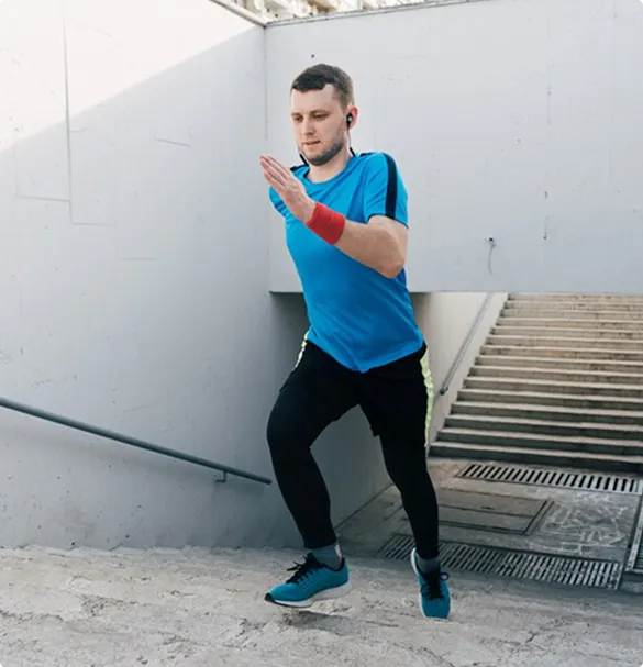 A man in athletic wear runs up concrete steps, wearing a blue shirt and black pants. Earbuds and a red wristband suggest focus and determination.