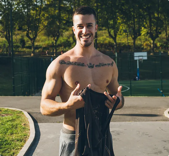 Smiling man outdoors, shirtless, holding tank top. Muscular build with chest tattoo. Background shows park with trees and a basketball court. Bright and cheerful mood.