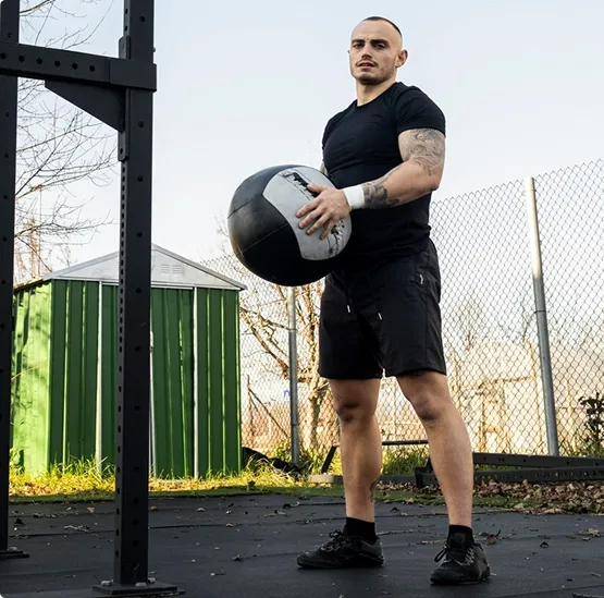 A man in black workout attire stands outdoors by a fence, holding a medicine ball. The setting is sunlit and conveys a focused, determined mood.