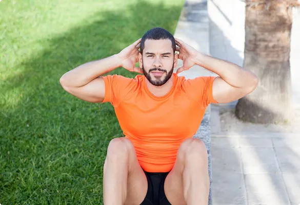A man in an orange shirt exercises outdoors, performing a sit-up on a grassy surface beside a stone path and tree, conveying focus and determination.