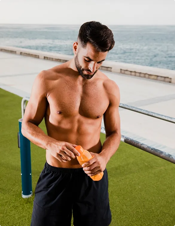 Shirtless man outdoors near the ocean, opening an orange bottle. He stands on a green artificial grass surface, wearing black shorts, evoking a fitness vibe.
