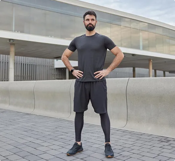 A man stands confidently outdoors, wearing a gray athletic outfit with leggings and sneakers. Behind him is a modern building with large windows.