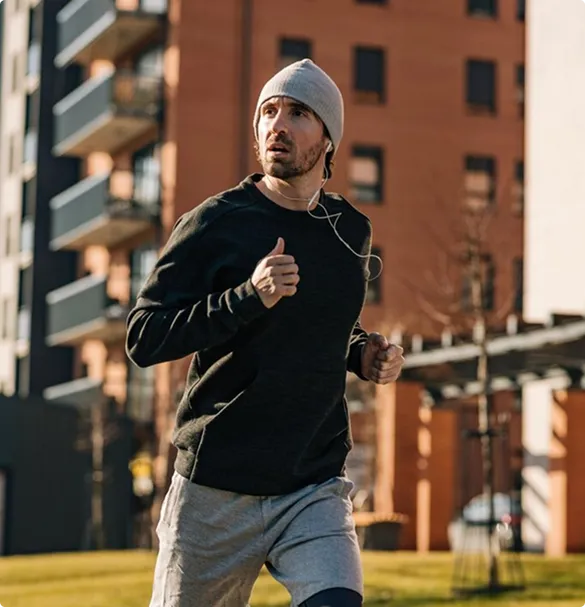 A man in a beanie jogs in a park, with greenery and pathways visible around him.