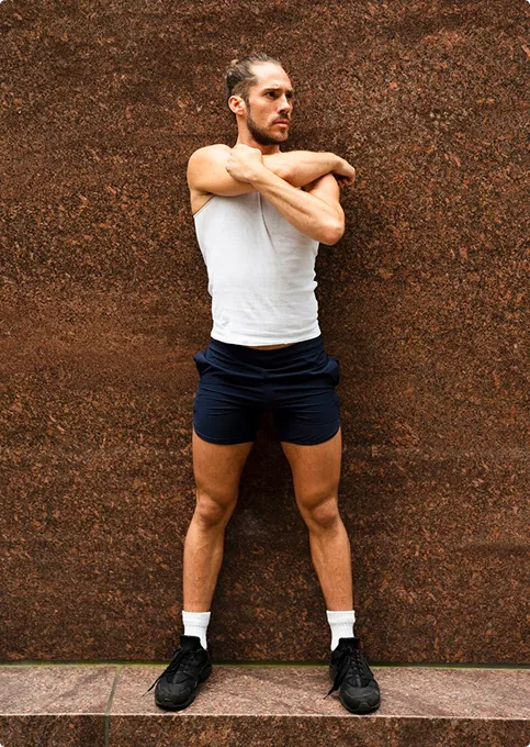 Man with tied-back hair stretches against a textured stone wall, wearing a white tank top, navy shorts, black sneakers, and white socks. He appears focused and athletic.
