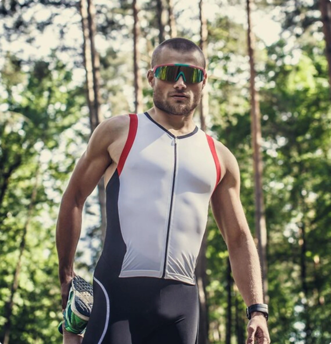 A man in a triathlon suit stands confidently among tall trees in a wooded area.